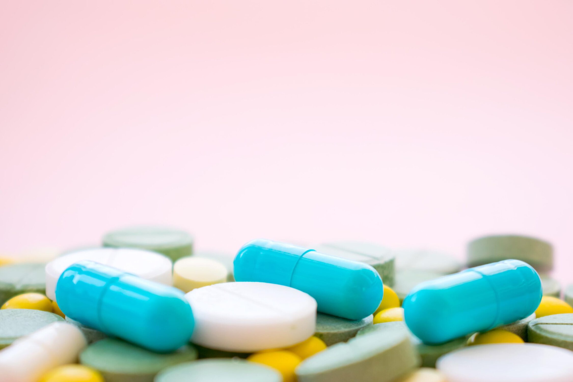 Blue and white prescription pills and capsules scattered on a surface with a soft pink background.