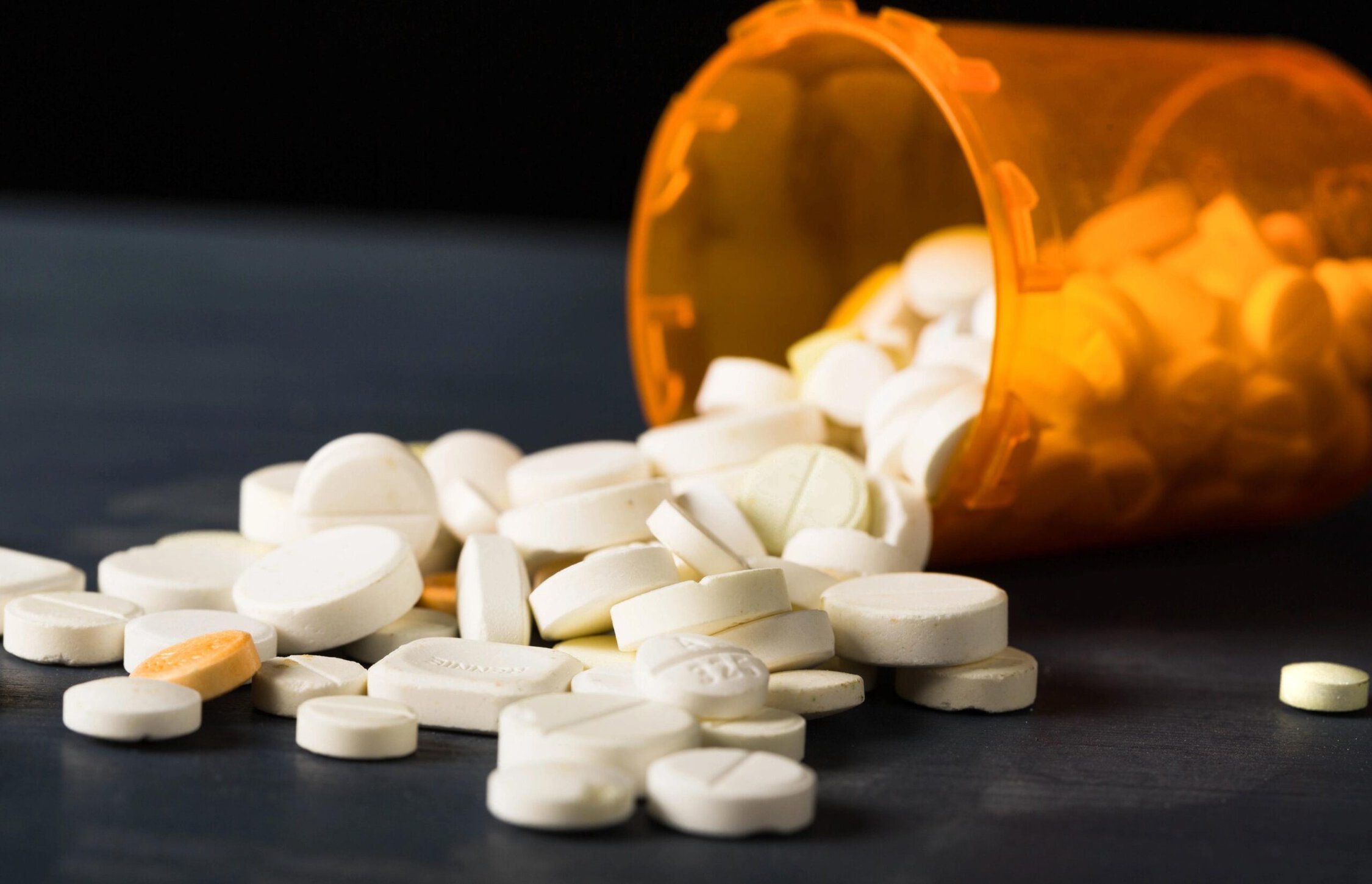 Opiate pills spilled from an orange prescription bottle on a dark surface.